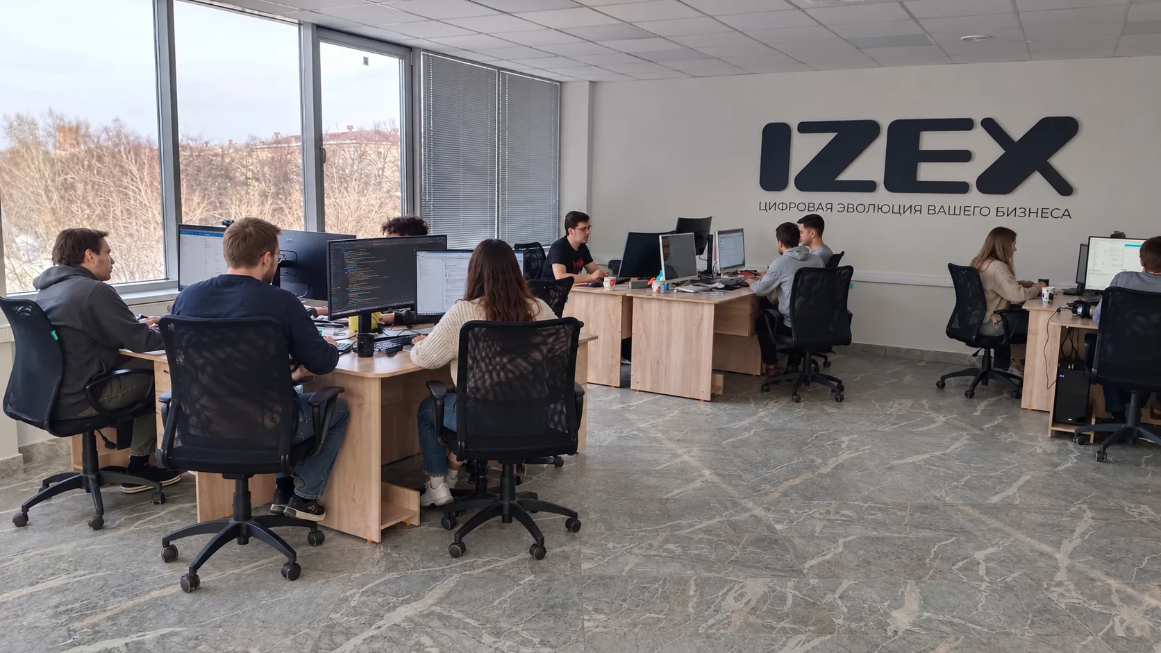 Сотрудники АЙЗЕКС за компьютерами в офисе с логотипом IZEX